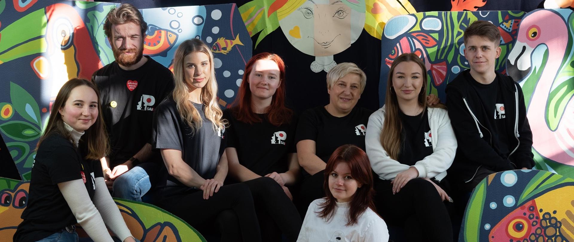  Zdjęcie przedstawia młodzież Studium. Wszyscy ubrani czarne ubrania z logiem Studium. Na środku dziewczyna robi szpagat.