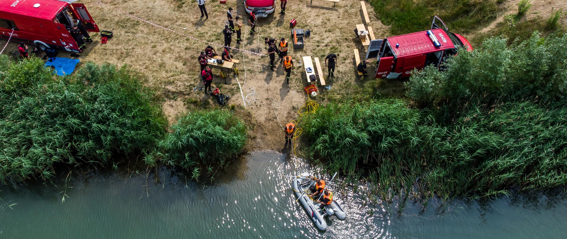 Na zdjęciu wykonane za pomocą drona (widok z góry) widoczne są trzy samochody, łódź ratownicza oraz strażacy ze Specjalistycznej Grupy Ratownictwa Wodno-Nurkowego "Nysa". 