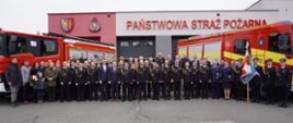Zdjęcie grupowe na tle dwóch samochodów pożarniczych oraz budynku komendy. Nad głowami napis Państwowa Straż Pożarna i logo PSP oraz herb Miasta Żory. 