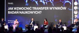 Pod niebieską ścianką z napisem „Jak wzmocnić transfer wyników badań naukowych?” w białych fotelach siedzi sześć osób, druga od lewej wiceminister Mrówczyńska w różowym garniturze.