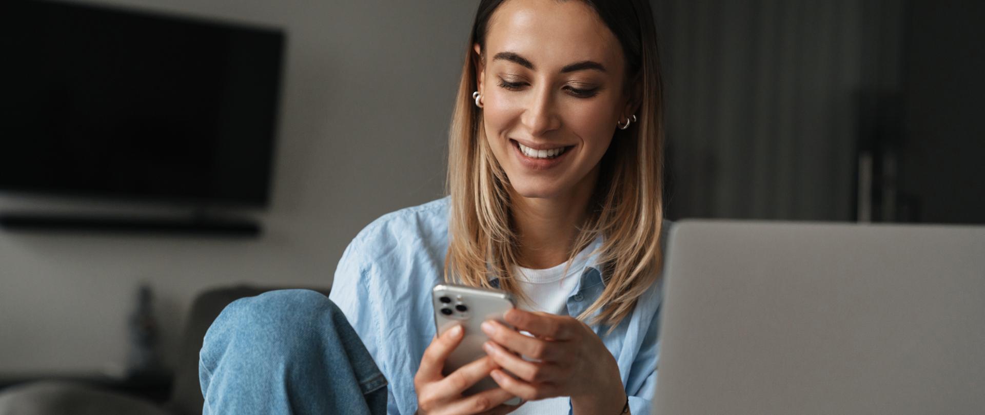 Uśmiechnięta kobieta z telefonem w rękach. Siedzi przy stole, na blacie stoi laptop.