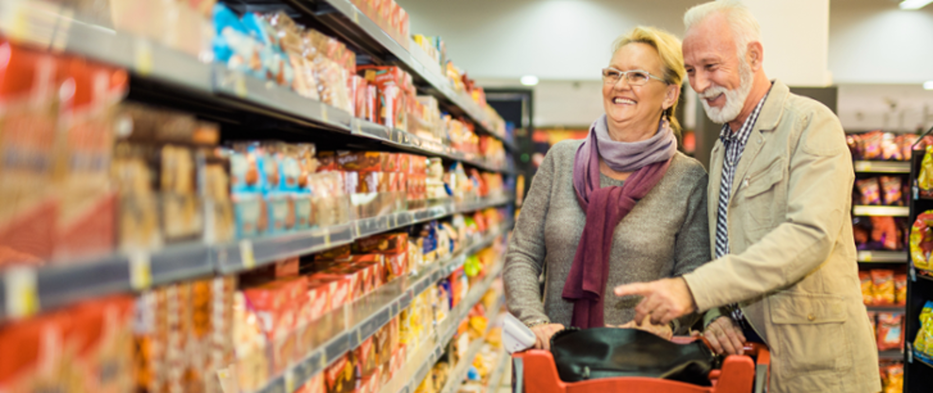 Małżeństwo seniorów robi zakupy w sklepie spożywczym; stoją przy półkach z produktami, rozmawiają i wybierają artykuły, trzymając koszyki na zakupy.