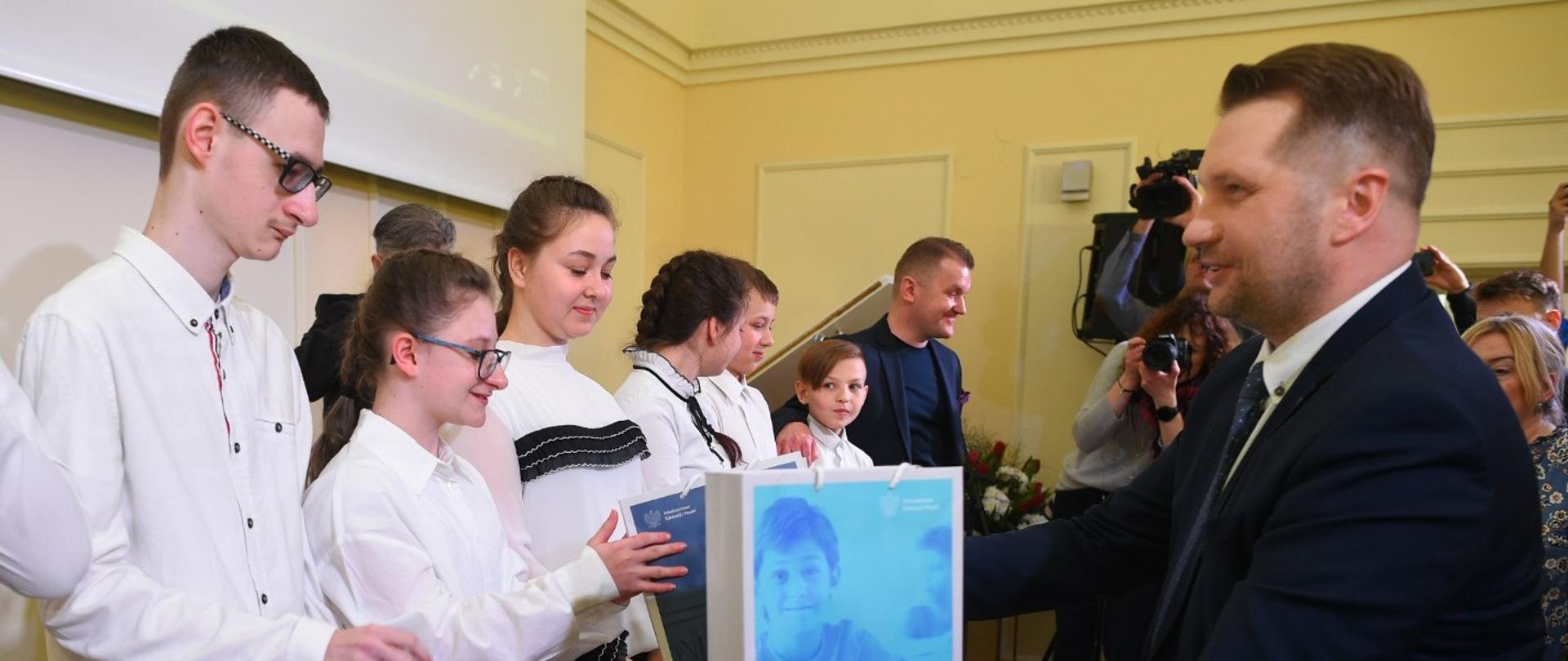 Grupa dzieci w białych bluzkach stoi pod ścianą przy tablicy. Z drugiej strony minister Przemysław Czarnek wręcza im upominek. W te grupa osób. 