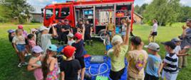 Zdjęcie przedstawia dzieci oraz wóz strażacki ze sprzętem podczas pokazu sprzętu na półkolonii dzieci