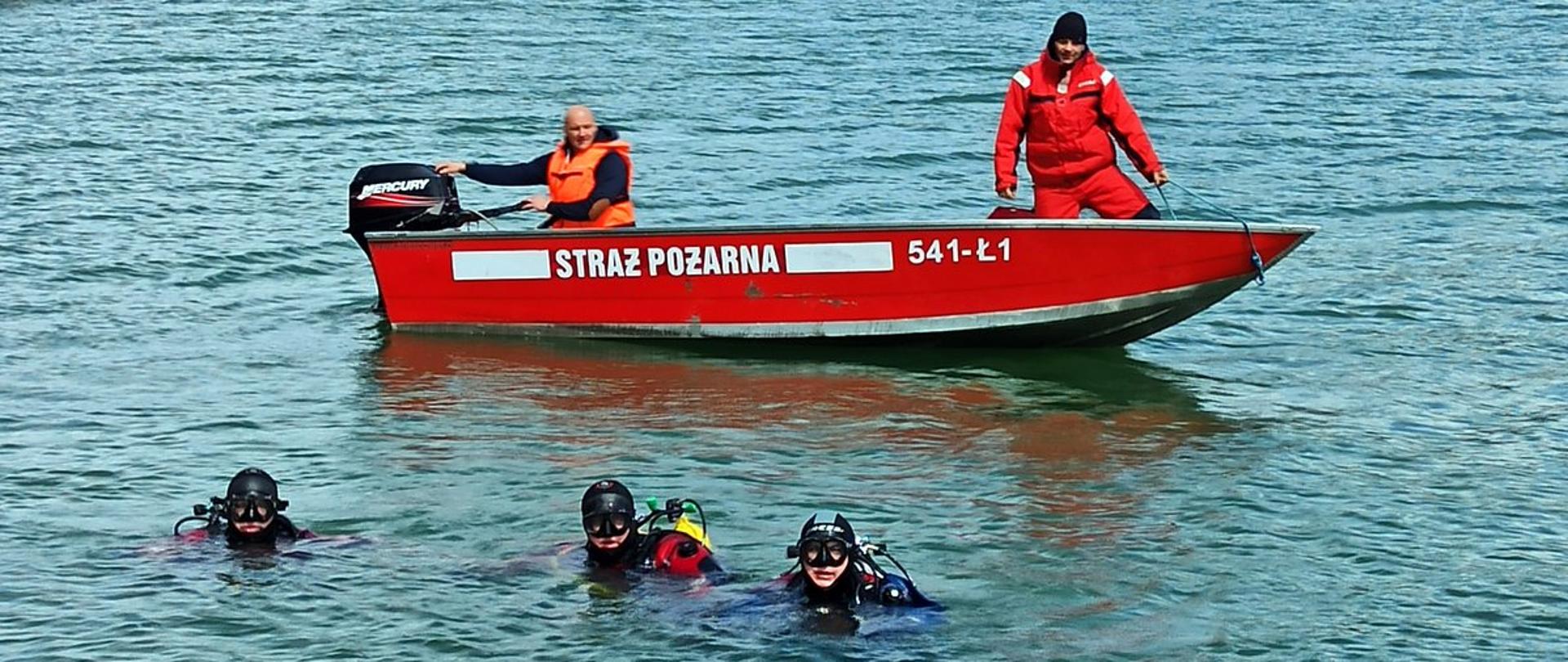 Na pierwszym planie widać trzech nurków w wodzie po nurkowaniu w tle łódź strażacką w jeziorze Solińskim, na której pokładzie znajduje sternik i ratownik wyposażeni w środki ochrony indywidualnej. 