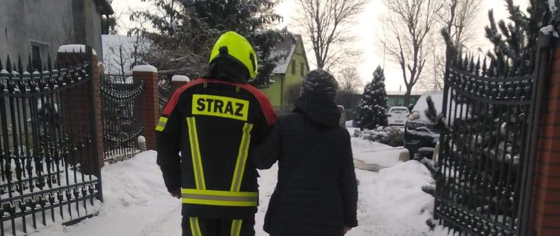 Zdjęcie przedstawia strażaka w umundurowaniu pomagającego kobiecie ubranej w czarną kurtkę w przemieszczeniu się.