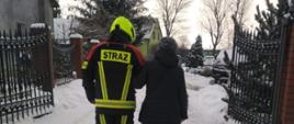 Zdjęcie przedstawia strażaka w umundurowaniu pomagającego kobiecie ubranej w czarną kurtkę w przemieszczeniu się.
