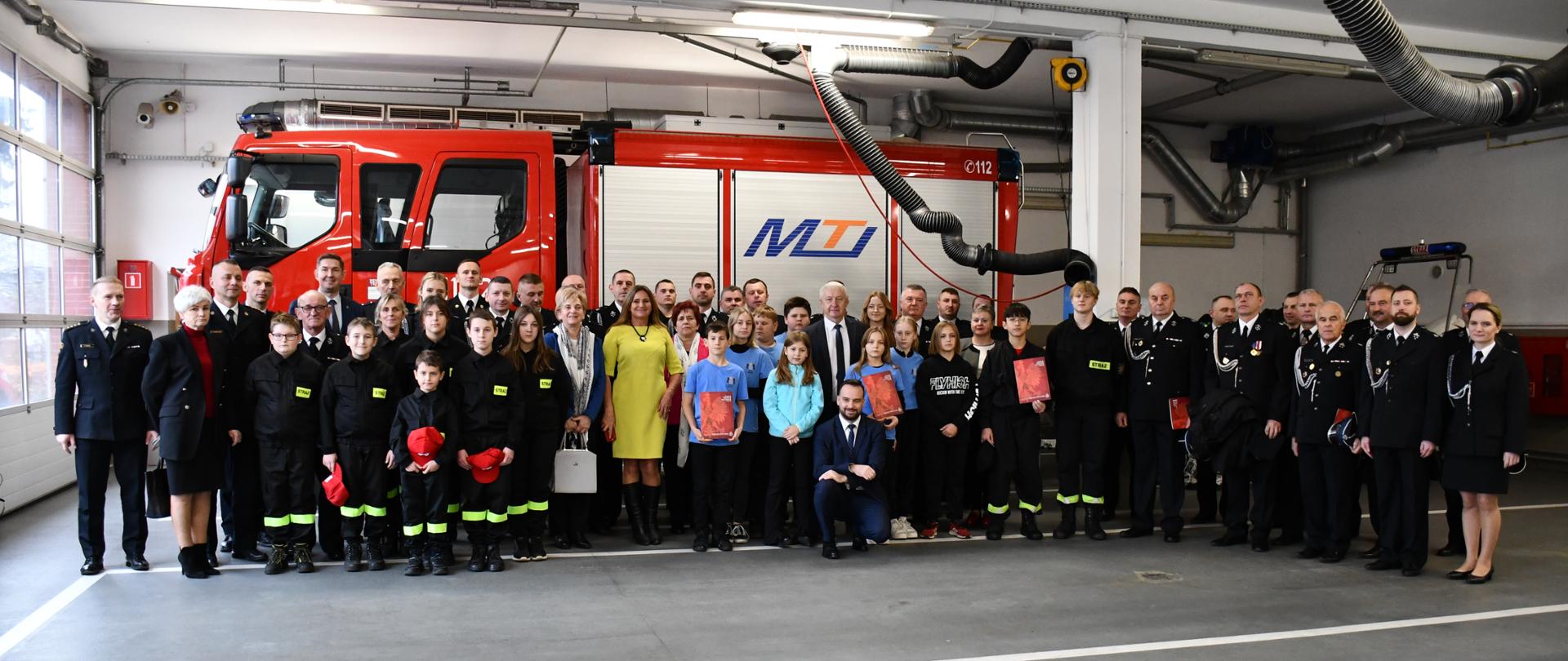 Uroczysta zbiórka z okazji wręczenia promes dla Młodzieżowych Drużyn Pożarniczych uczestnicy uroczystości na tle samochodu pożarniczego. 