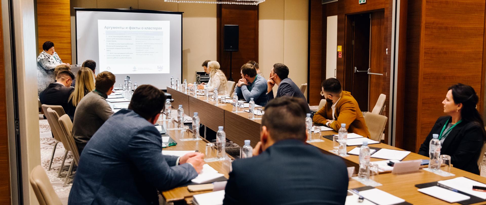 Grupa osób siedzi przy stole konferencyjnym i obserwuje prezentację 