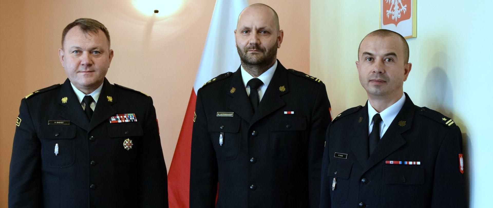 Zdjęcie zrobione wewnątrz pomieszczenia. Na zdjęciu trzech oficerów Państwowej Straży Pożarnej w mundurach wyjściowych.