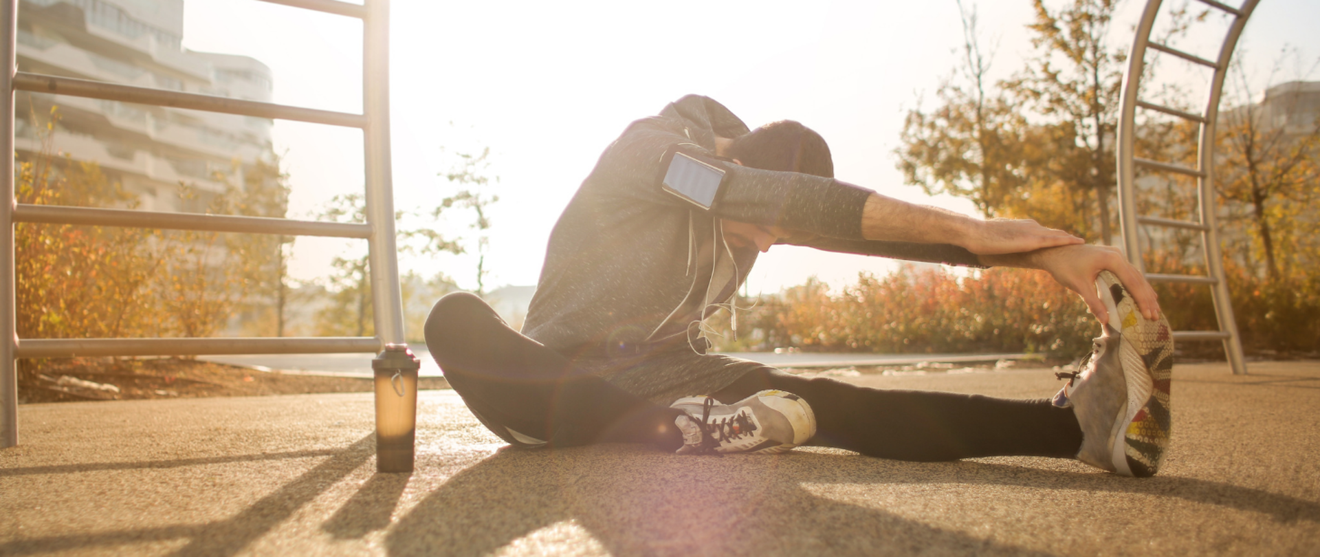 Młody mężczyzna w stroju sportowym rozciąga na siłowni przy ulicy. Widać prosty sprzęt gimnastyczny, bidon, jesienne drzewa i miejskie zabudowanie
