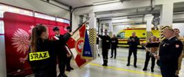 Uroczystość ślubowania strażaków w garażu Komendy Powiatowej Państwowej Straży Pożarnej w Obornikach. Funkcjonariusze w piaskowych ubraniach służbowych stoją w dwóch szeregach naprzeciwko siebie, tworząc przejście prowadzące do miejsca ceremonii. W głębi zdjęcia, na tle czerwonego baneru PSP, odbywa się uroczyste ślubowanie.