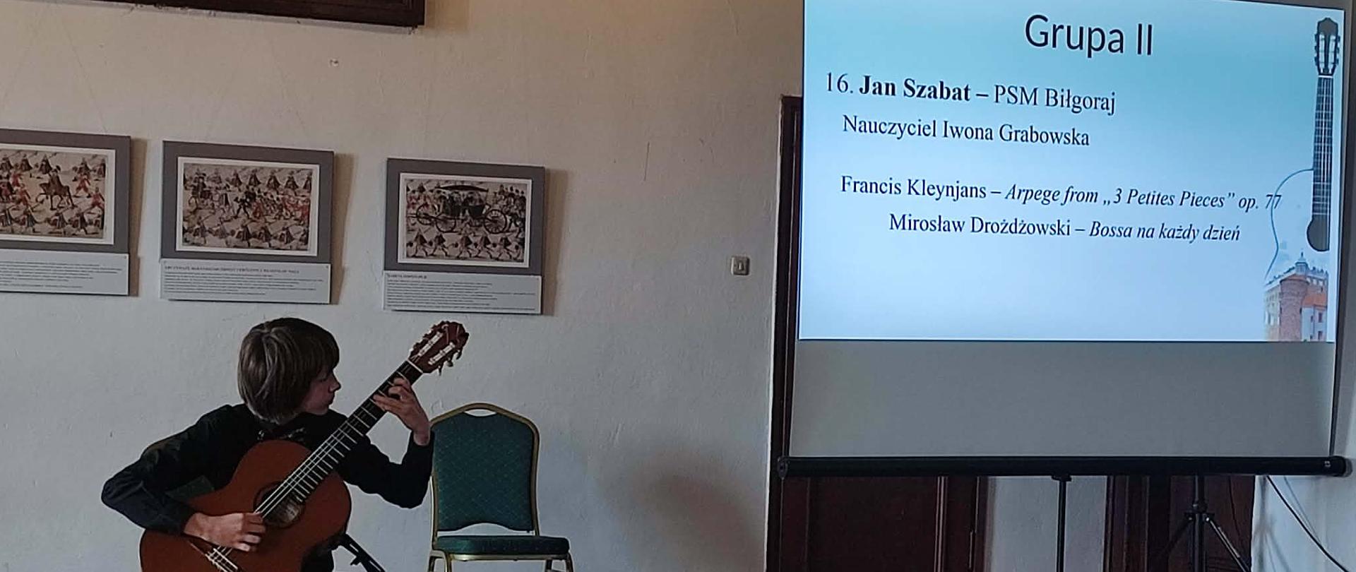 Na zdjęciu widać Jana Szabata podczas występu w sali konkursowej na zamku. Chłopiec siedzi na krześle i gra na gitarze klasycznej, skupiony na wykonaniu utworu. Obok znajduje się ekran projekcyjny z informacjami o uczestniku – „Jan Szabat – PSM Biłgoraj”, nauczycielce Iwonie Grabowskiej oraz wykonywanych utworach Francisa Kleynjansa i Mirosława Drożdżowskiego. Wnętrze ma kameralny, elegancki charakter – drewniana podłoga, jasne ściany. Zdjęcie oddaje moment pełnego skupienia i artystycznego zaangażowania młodego muzyka podczas występu konkursowego.