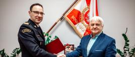 Uścisk dłoni między funkcjonariuszem Państwowej Straży Pożarnej a mężczyzna w garniturze. W tle sztandar PSP, trąbka hełmowa oraz elementy dekoracyjne gabinetu.
