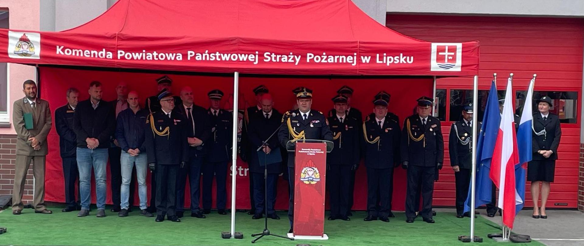 Zdjęcie przedstawia uroczystość oficjalną odbywającą się przed budynkiem straży pożarnej. W centralnej części obrazu znajduje się czerwony namiot ekspozycyjny z białym napisem: "Komenda Powiatowa Państwowej Straży Pożarnej w Lipsku". Pod namiotem, na zielonej wykładzinie imitującej trawę, stoi grupa osób – w większości są to funkcjonariusze straży pożarnej w galowych mundurach z odznaczeniami oraz cywile w strojach wizytowych.
Po prawej stronie, przy czerwonej mównicy z logo straży, stoi jeden z funkcjonariuszy przemawiający do zgromadzonych. Obok namiotu, po prawej stronie, widoczne są dwie flagi na masztach: flaga Unii Europejskiej oraz flaga Polski. Tło stanowi szara elewacja budynku z czerwonymi bramami garażowymi. Całość sceny odbywa się na placu wyłożonym szarą kostką brukową.