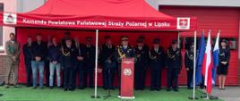 Zdjęcie przedstawia uroczystość oficjalną odbywającą się przed budynkiem straży pożarnej. W centralnej części obrazu znajduje się czerwony namiot ekspozycyjny z białym napisem: "Komenda Powiatowa Państwowej Straży Pożarnej w Lipsku". Pod namiotem, na zielonej wykładzinie imitującej trawę, stoi grupa osób – w większości są to funkcjonariusze straży pożarnej w galowych mundurach z odznaczeniami oraz cywile w strojach wizytowych.
Po prawej stronie, przy czerwonej mównicy z logo straży, stoi jeden z funkcjonariuszy przemawiający do zgromadzonych. Obok namiotu, po prawej stronie, widoczne są dwie flagi na masztach: flaga Unii Europejskiej oraz flaga Polski. Tło stanowi szara elewacja budynku z czerwonymi bramami garażowymi. Całość sceny odbywa się na placu wyłożonym szarą kostką brukową.