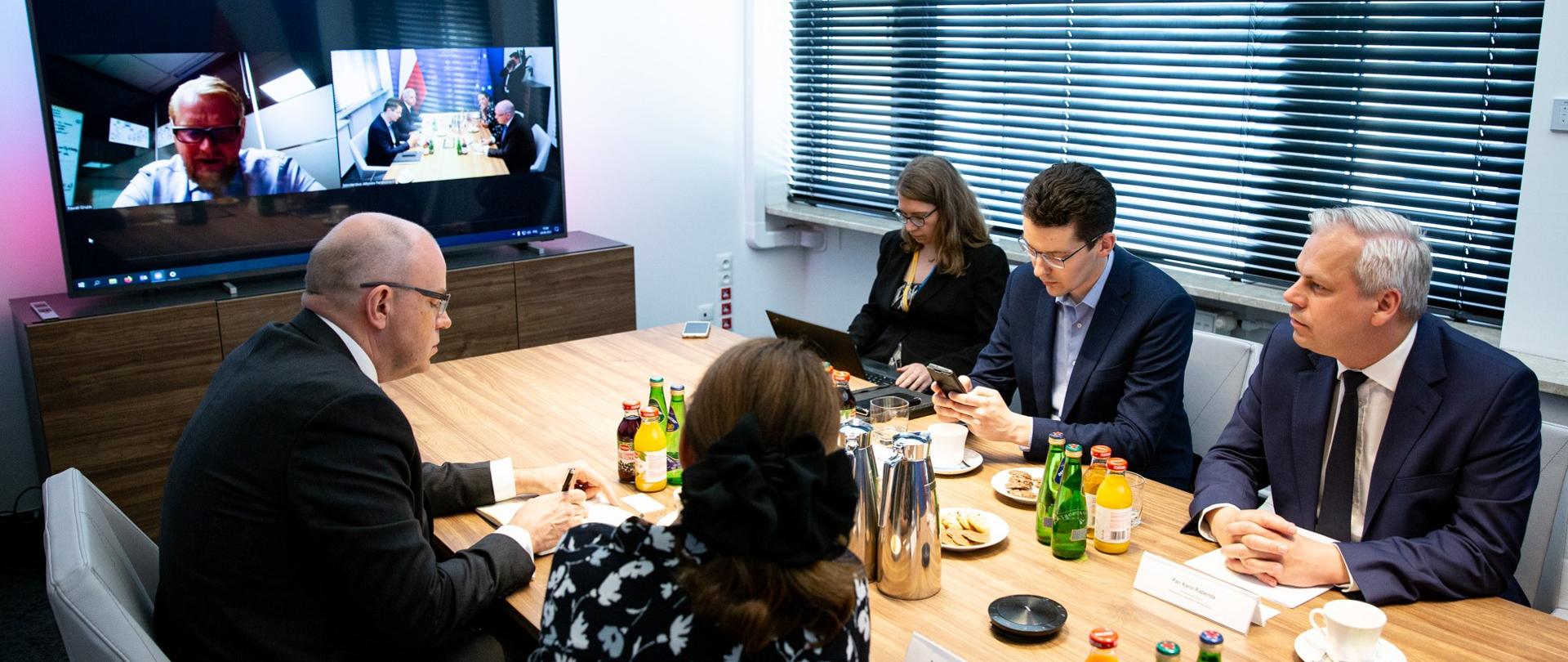 Przy stole siedzi pięć osób w tym jedna obrócona tyłem, jedna bokiem. W tle ekran z połączeniem online spotkania. Na stole stoją butelki z wodą, soki, filiżanki.