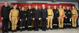 Grupa kilkunastu strażaków w umundurowaniu galowym oraz bojowym pozuje do wspólnego zdjęcia wewnątrz budynku. W tle widoczny jest czerwony baner Państwowej Straży Pożarnej. Kilka osób trzyma czerwone teczki, a cała grupa stoi w równym szeregu i uśmiecha się do kamery.