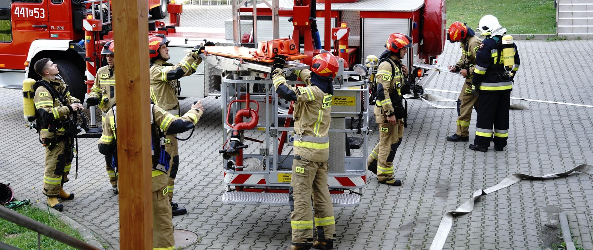 Na zdjęciu widoczni strażacy w ubraniach specjalnych koloru piaskowego w hełmach strażackich i sprzęt pożarniczy w tym w tle podnośnik hydrauliczny podczas ćwiczeń na szpitalu w Gorlicach.