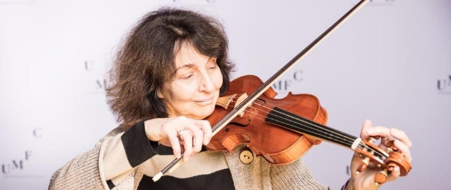 Na zdjęciu widoczna jest kobieta w średnim wieku, grająca na skrzypcach. Ma ciemne, krótkie włosy i jest ubrana w beżowo-czarny sweter w pasy. Skupiona na grze, delikatnie trzyma smyczek i przykłada instrument do szyi, demonstrując technikę gry. Tło jest neutralne, jasne, z nieczytelnym napisem.