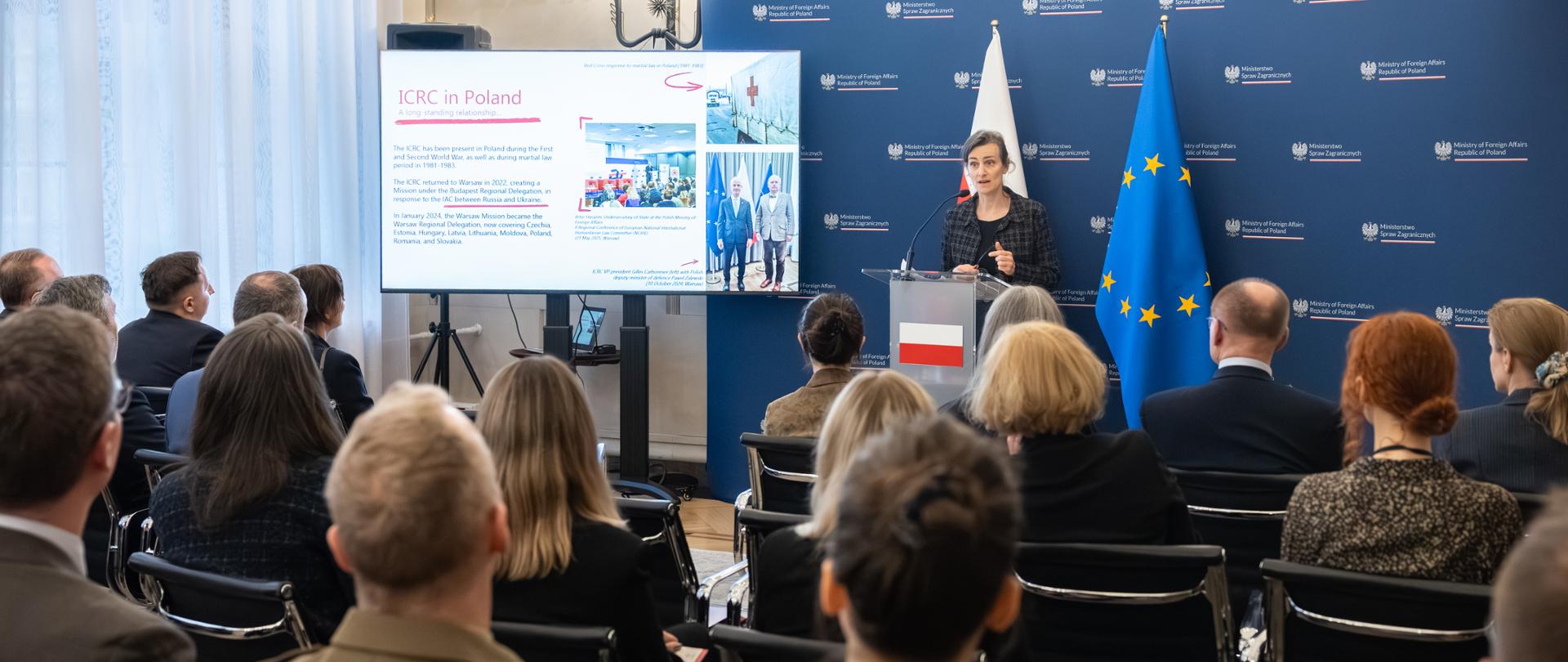 The photo shows a group of people. One woman is standing at the podium. Behind her are the Polish and EU flags. In front of her are a dozen or so people sitting in chairs and facing her. 