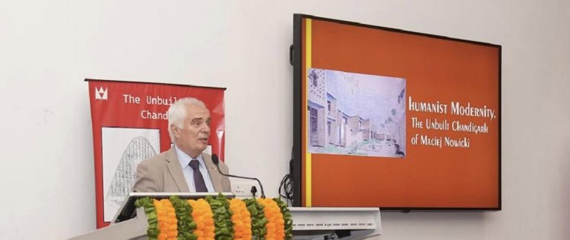 Kierownik Placówki dr Piotr Antoni Świtalski, przemawia przy mównicy ozdobionej girlandami z pomarańczowych i zielonych kwiatów. Na ekranie obok wyświetlony jest tytuł wystawy „Humanist Modernity: The Unbuilt Chandigarh of Maciej Nowicki” oraz reprodukcja rysunku architektonicznego.