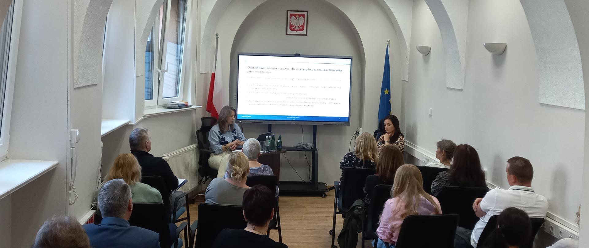 Pracownicy Wojewódzkiego Inspektoratu Inspekcji Handlowej na szkoleniu pt. "Prewencja zagrożeń psychospołecznych w miejscu pracy." Pracownicy siedzą na czarnych krzesłach naprzeciwko dwóch kobiet - prelegentek. Pomiędzy prelegentkami znajduje się monitor multimedialny, na którym wyświetla się prezentacja. Powyżej monitora znajduje się godło Polski. 