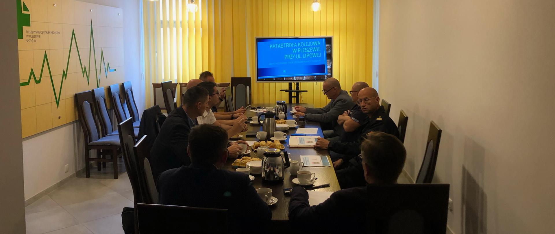 Grupa mężczyzn siedzi przy stole w zaciemnionym pokoju, w tle widać ekran telewizyjny z wyświetlaną prezentacją