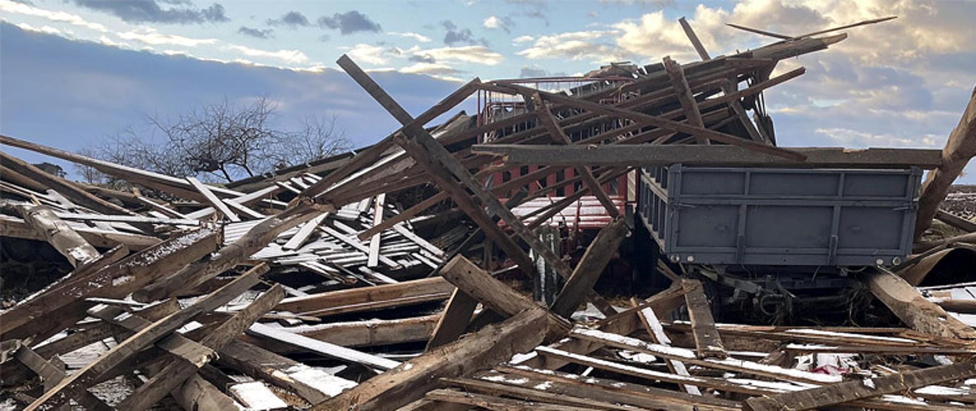 Na środku zdjęcia dwie przyczepy zasypane stertą desek, dookoła leżą porozrzucane deski przez silne wiatry, niebo ciemne, burzowe z miejscowymi przejaśnieniami