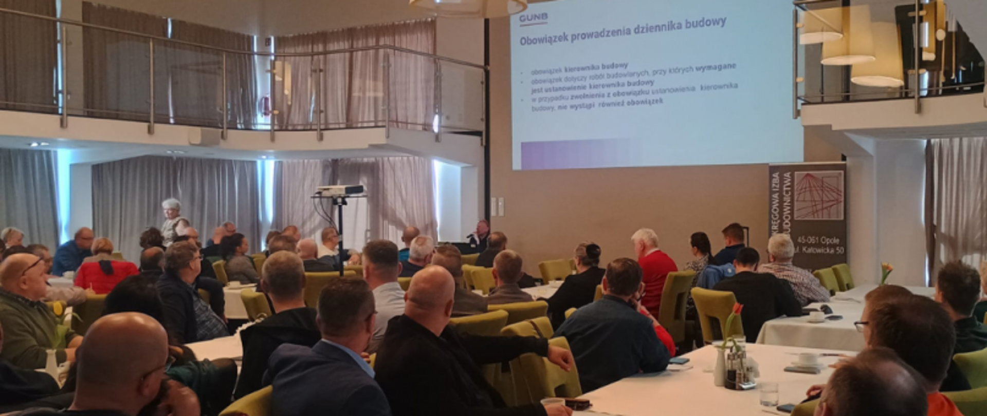 Szkolenie organizowane przez Główny Urząd Nadzoru Budowlanego (GUNB) z zakresu Elektronicznego Dziennika Budowy (EDC) i Centralnego Katalogu Obiektów Budowlanych (c-KOB) dla przedstawicieli Opolskiej Okręgowej Izby Inżynierów Budownictwa; uczestnicy siedzą przy stołach, na ekranie wyświetlona jest prezentacja.