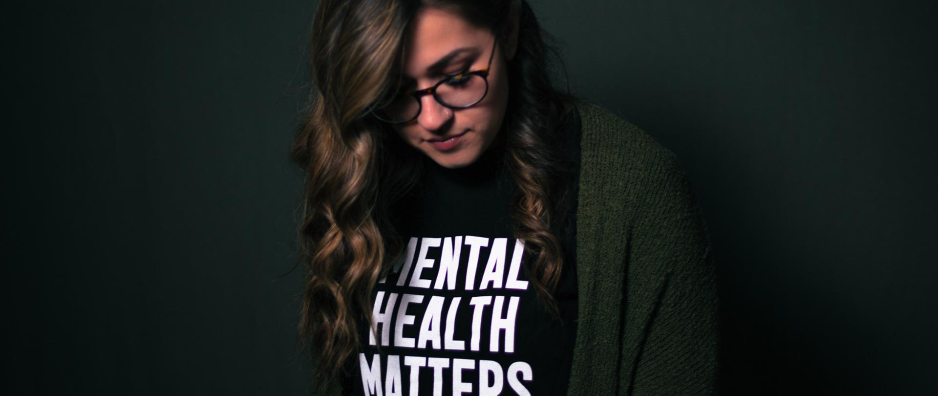 ciemnowłosa kobieta stojąca ze spuszczoną głową na tle ciemnej ściany ubrana w czarny podkoszulek z napisem w języku angielskim 