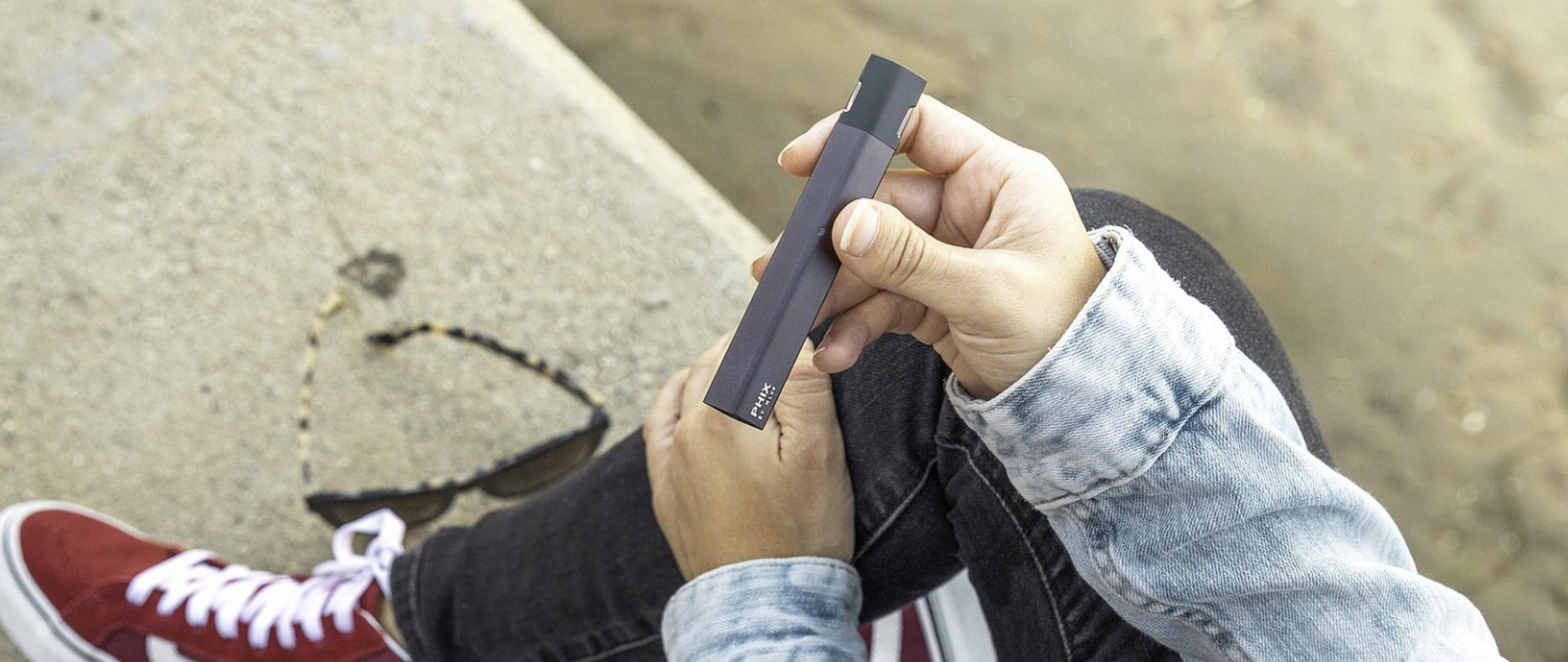 Grafika ukazuje osobę trzymającą e-papierosa i siedzącą na murku.