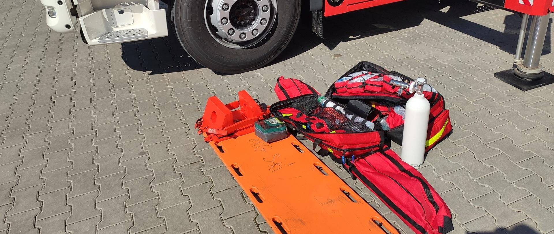 Zdjęcie obrazuje nosze typu deska ortopedyczna walizkę medyczną, szyny do stabilizacji, butlę tlenową, część samochodu pożarniczego