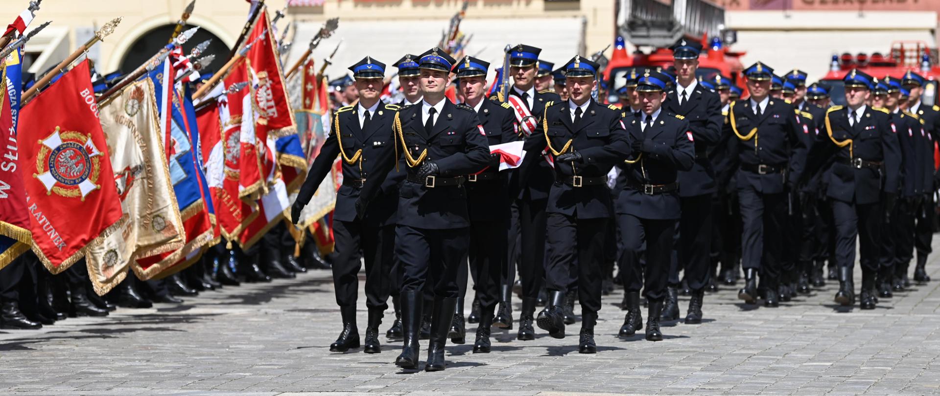 Na zdjęciu widać kompanię reprezentacyjną defilującą podczas uroczystości Dnia Strażaka. Po lewej stronie stoją poczty sztandarowe straży pożarnych.