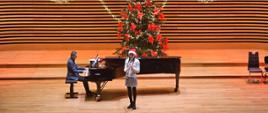 Dziewczynka gra na klarnecie, za nią na fortepianie gra mężczyzna. Z tyłu za fortepianem stoi udekorowana choinka.