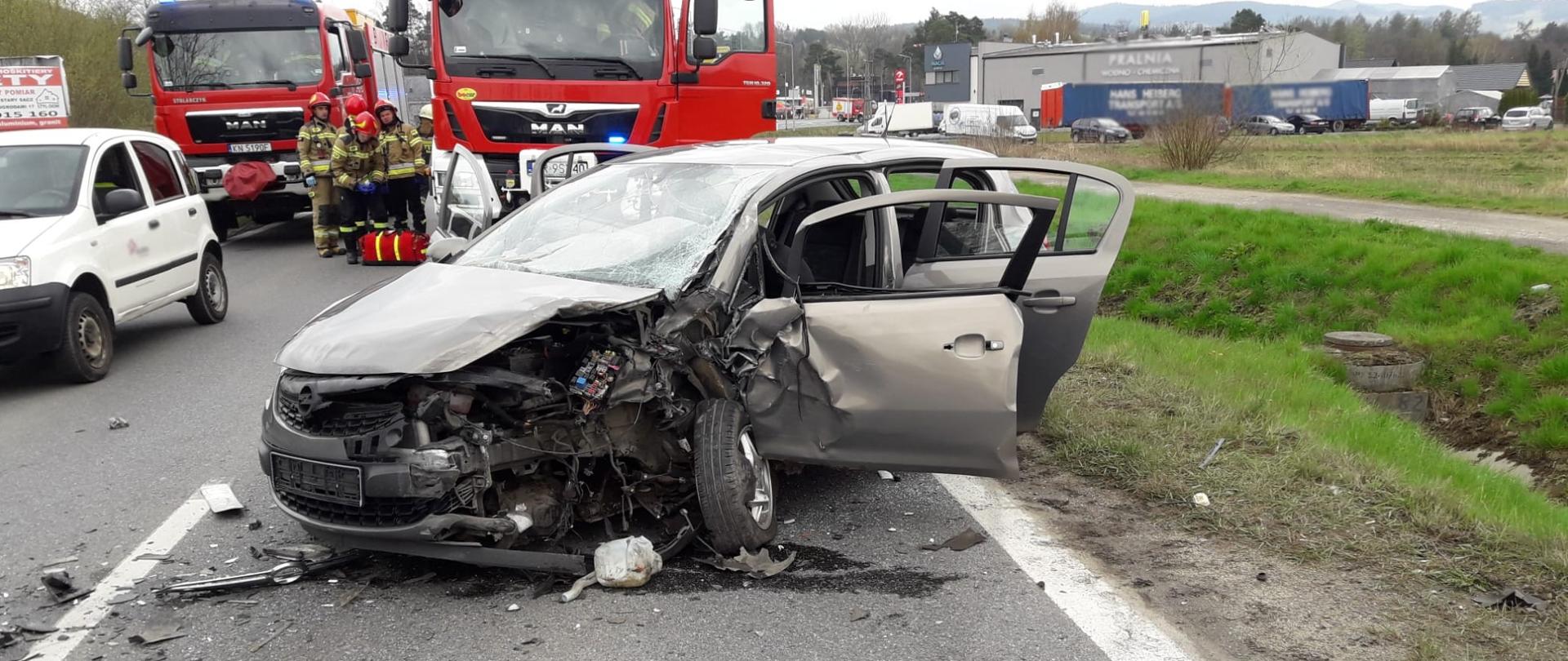 Rozbity samochód osobowy a za nim samochody pożarnicze.