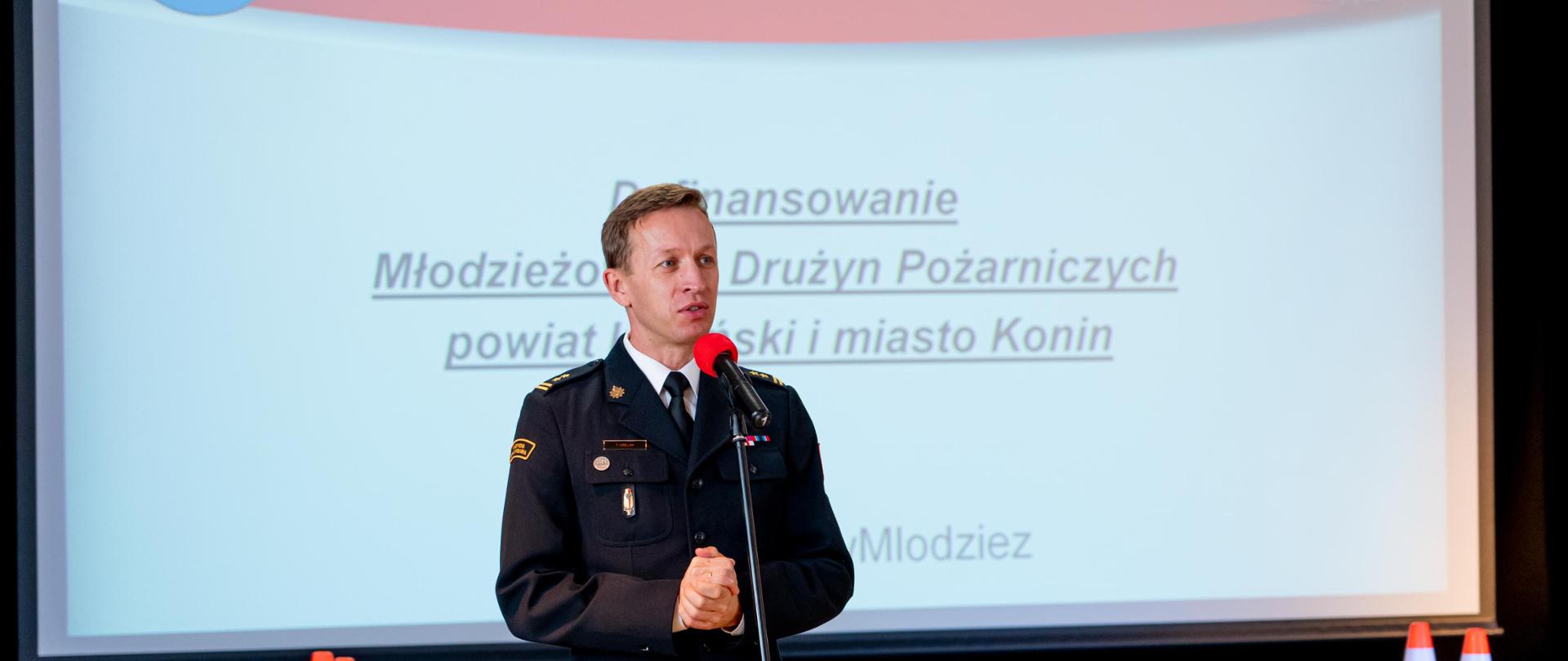 strażak stoi przed mikrofonen, za nim duży ekran z napisem : gmina kaizmierz biskupi , państwowa straż pożarna, poniżej napis: dofinansowanie młodzieżowych drużyn pożarniczych powiat koniński i miasto konin