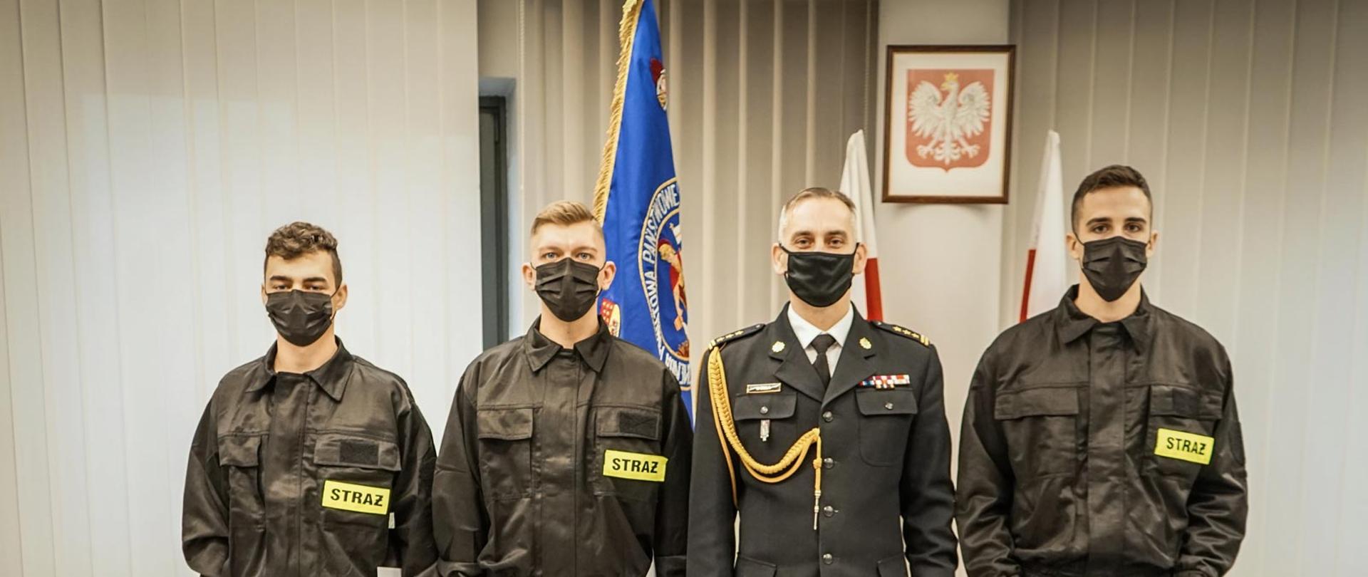 Trzech strażaków ubranych w ubraniach koszarowe oraz komendant PSP w Lublińcu w mundurze wyjściowym. W tle sztandar jednostki oraz godło państwowe