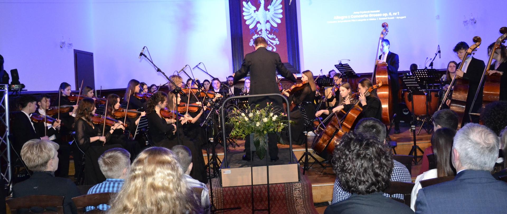 zdjęcie przedstawia orkiestrę symfoniczną naszej szkoły podczas koncertu w sali królewskiej, w tle na białej ścianie zawieszone jest wiecie godło polski