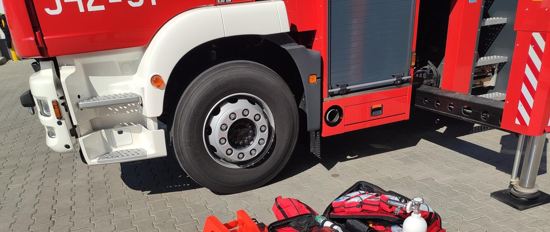 Zdjęcie obrazuje nosze typu deska ortopedyczna walizkę medyczną, szyny do stabilizacji, butlę tlenową, część samochodu pożarniczego