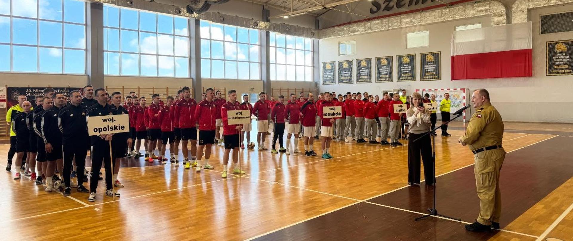 Uczestnicy turnieju stoją w rzędach na hali sportowej z tabliczkami województw, podczas oficjalnego otwarcia wydarzenia prowadzonego przez strażaka PSP.