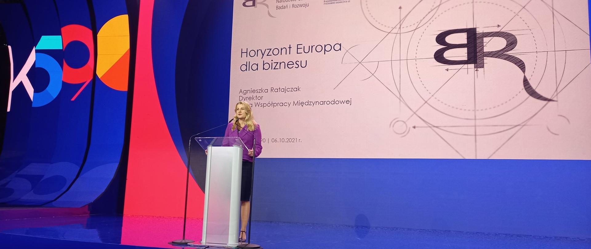 Widok na scenę. Na scenie przemawiająca dyrektor Agnieszka Ratajczak. Za nią ekran, na nim wyświetlony slajd z napisem: NCBR, KPK, Horyzont Europa dla biznesu, Agnieszka Ratajczak, dyrektor Biura Współpracy Międzynarodowej NCBR