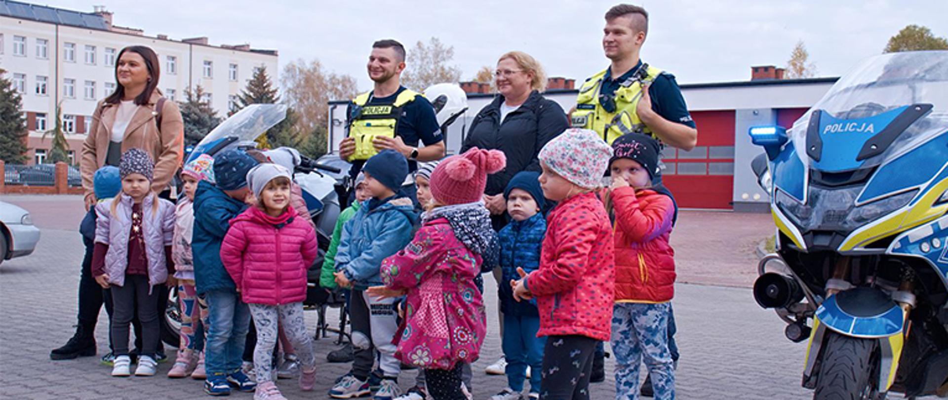 grupka dzieci wraz z opiekunkami i policjantami pozują do pamiątkowego zdjęcia z motorami za nimi znajdują się garaże na samochody strażackie