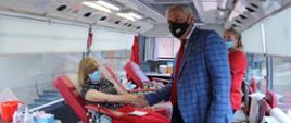 Na zdjęciu wojewoda opolski stojący w centralnej części kadru. Stoi bokiem do kadru. Trzyma za dłoń, siedzącą na fotelu medycznym, pracownicę urzędu, która oddaje krew. Za wojewodą w tle widać pielęgniarkę, ubraną w czerwony fartuszek. Zdjęcie wykonano w krwiobusie. Pracownica oddająca krew, spogląda nieco w dół. Obok niej widać aparaturę, pobierającą krew. 