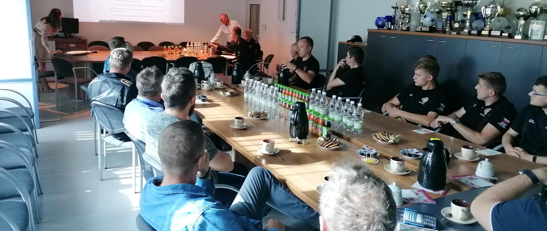 Całość odbywa się na sali wideokonferencyjnej. Wszyscy uczestnicy siedzą przy stole, w tle prezentacja.