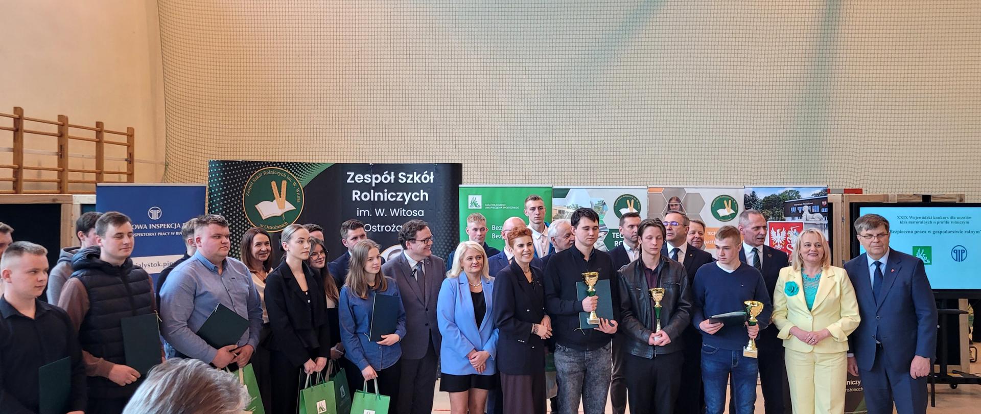 Grupa nagrodzonych uczestników konkursu wraz z opiekunami i członkami jury pozuje do zdjęcia na tle banerów różnych instytucji.