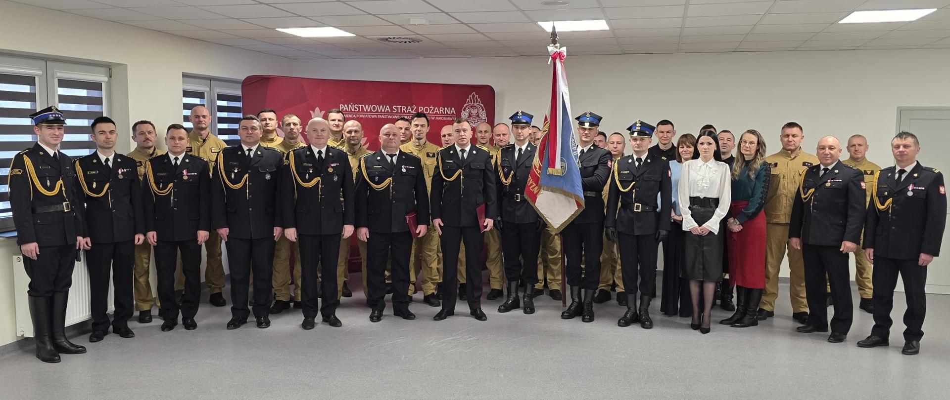 Grupa funkcjonariuszy Państwowej Straży Pożarnej w umundurowaniu służbowym i galowym, ustawionych w kilku rzędach do wspólnego zdjęcia, na tle ścianki z napisem „Państwowa Straż Pożarna