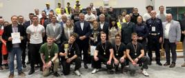 Grupa uczestników i organizatorów pozuje do wspólnego zdjęcia, część osób trzyma puchary, medale i dyplomy.