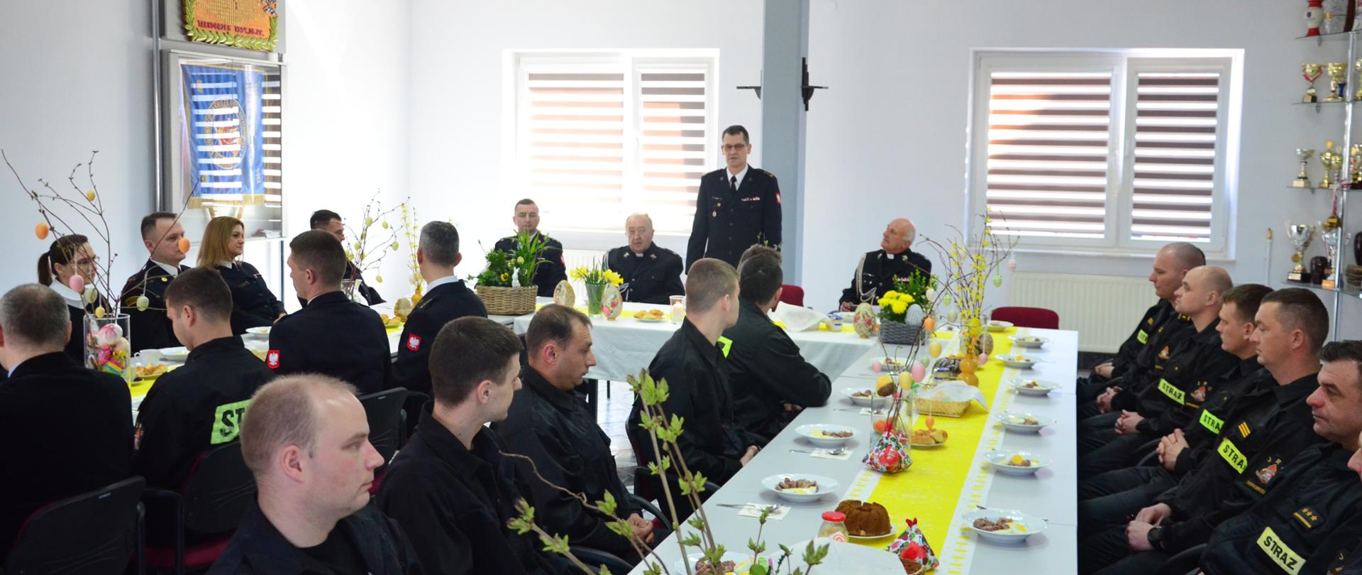 Przy świątecznie przyzdobionym stole, w dużym pomieszczeniu, siedzą strażacy. Jeden ze strażaków przemawia stojąc. 