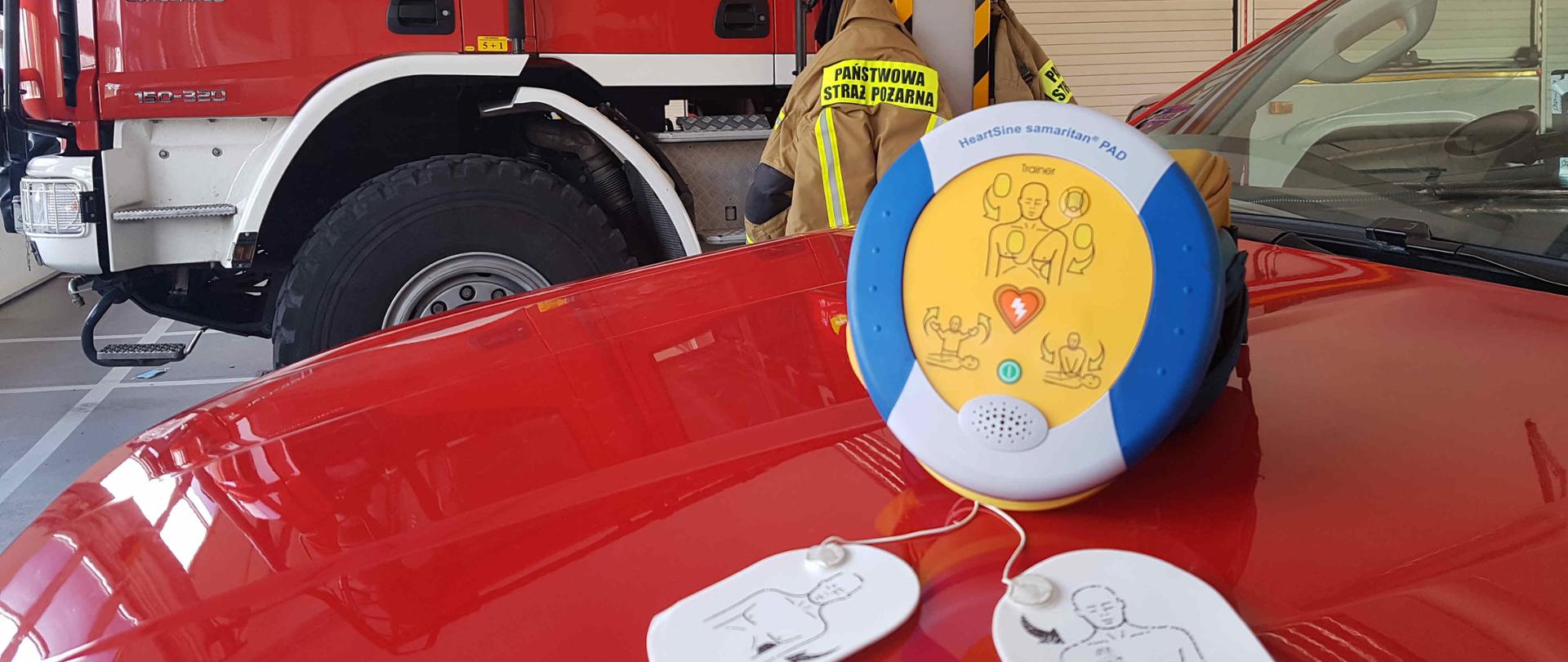 Zdjęcie zrobione na w garażu JRG Sandomierz. Na zdjęciu widać samochody strażackie, ubrania i hełmy wiszące na wieszakach. Na jednym z samochodów na masce lezy urządzenie AED. 